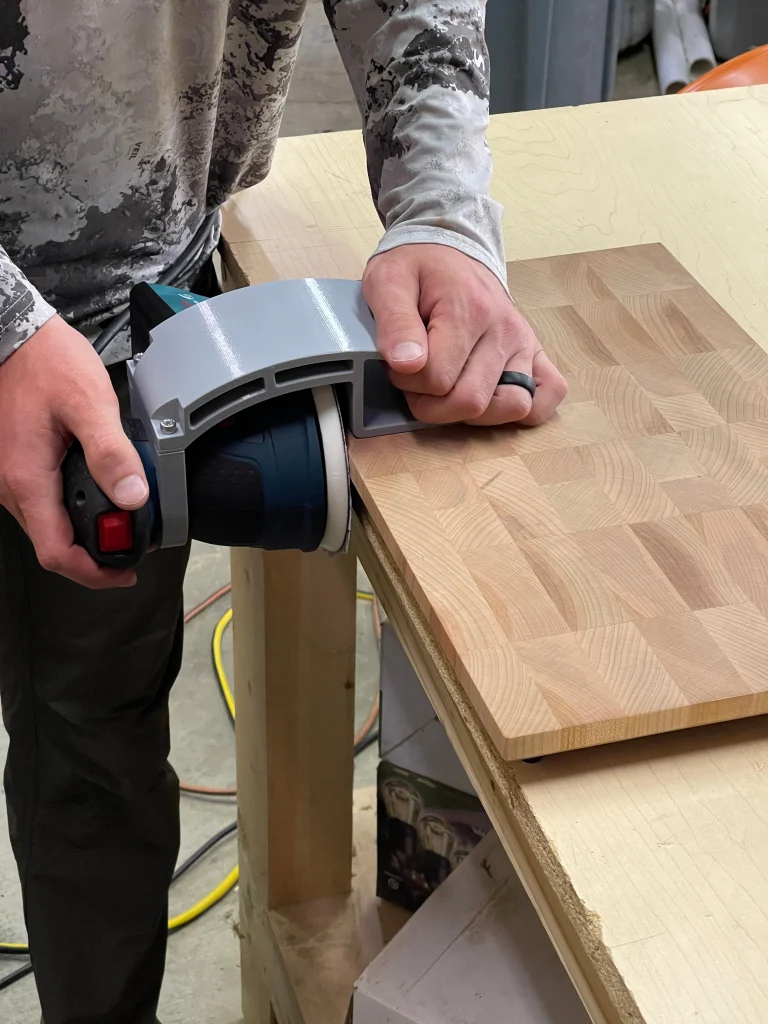 Hands guiding a Bosch orbital sander with an Xact90 guide along the edge of a wooden board